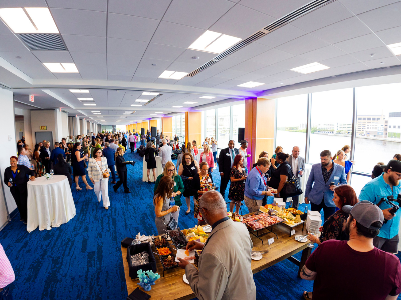 waterfront meeting room with floor to ceiling windows looking out over the water filled with attendees in business casual attire with reception tables and a full table of charcuterie.