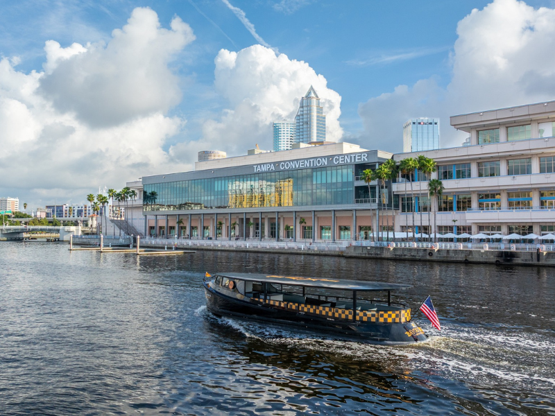 Exterior of modernized Tampa Convention Center façade made up of a glass and silver structure over the Tampa Riverwalk and water with blue skies in the background.
