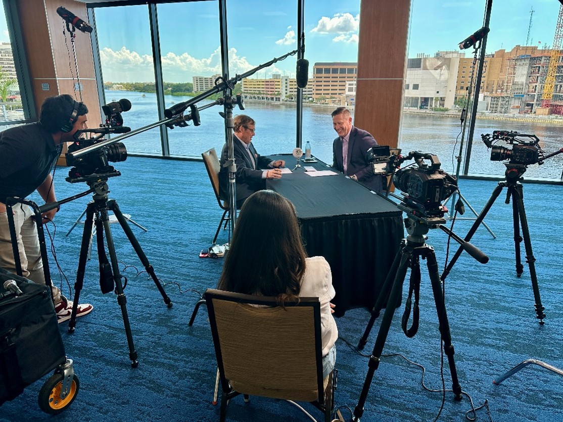 David Ingram sits down at table for podcast conversation with cameras and microphones in waterfront meeting room.