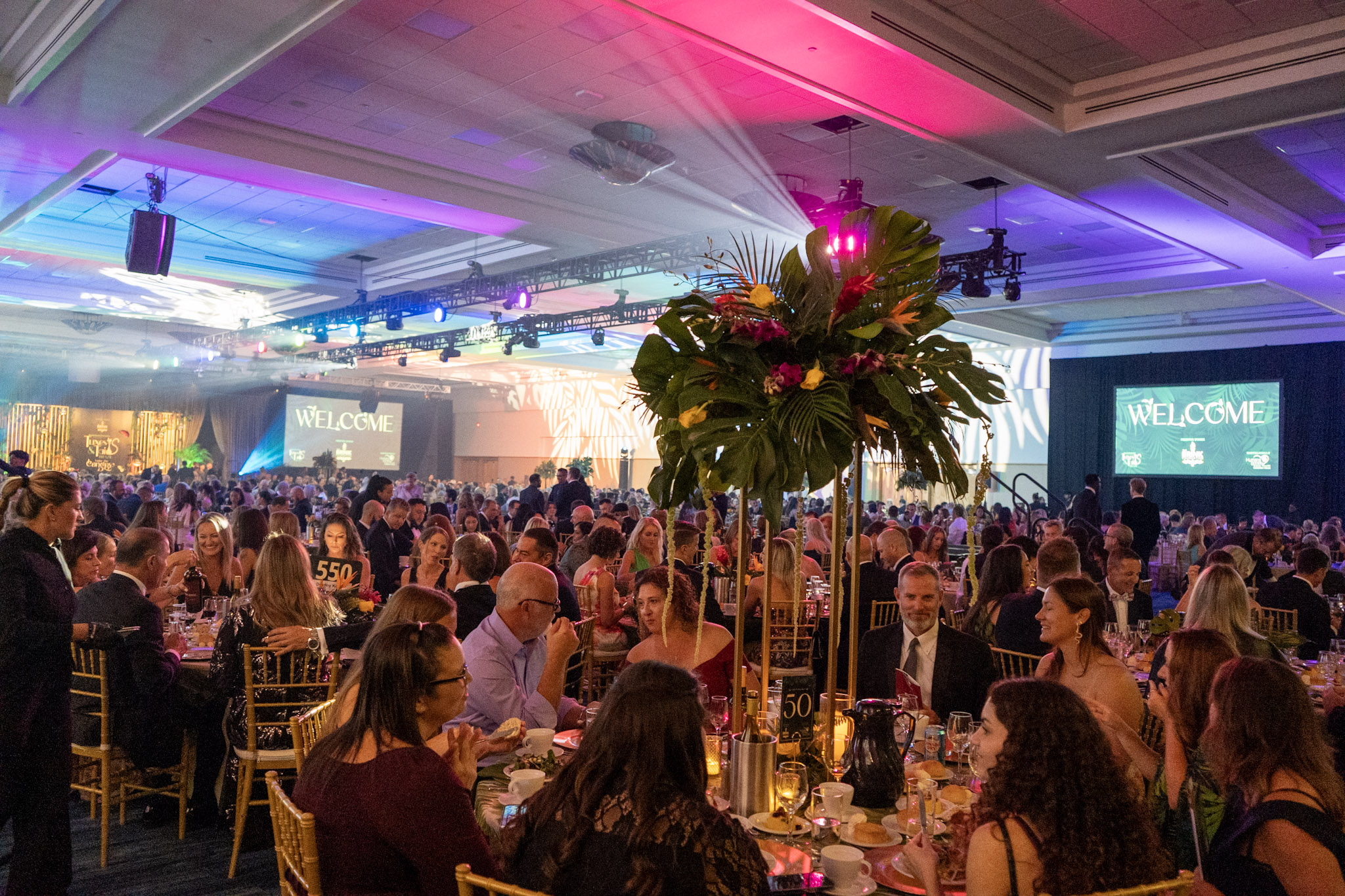 Ballroom inside Tampa Convention Center during gala with tables full of attendees in formal wear, colorful lights, and large floral tabletop displays.