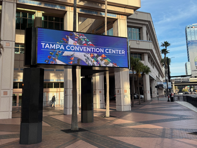 A modern digital electronic marquee sign standing on a paved walkway in front of the Tampa Convention Center building, displaying a blue background graphic with a sailboat and the text "TAMPA CONVENTION CENTER."