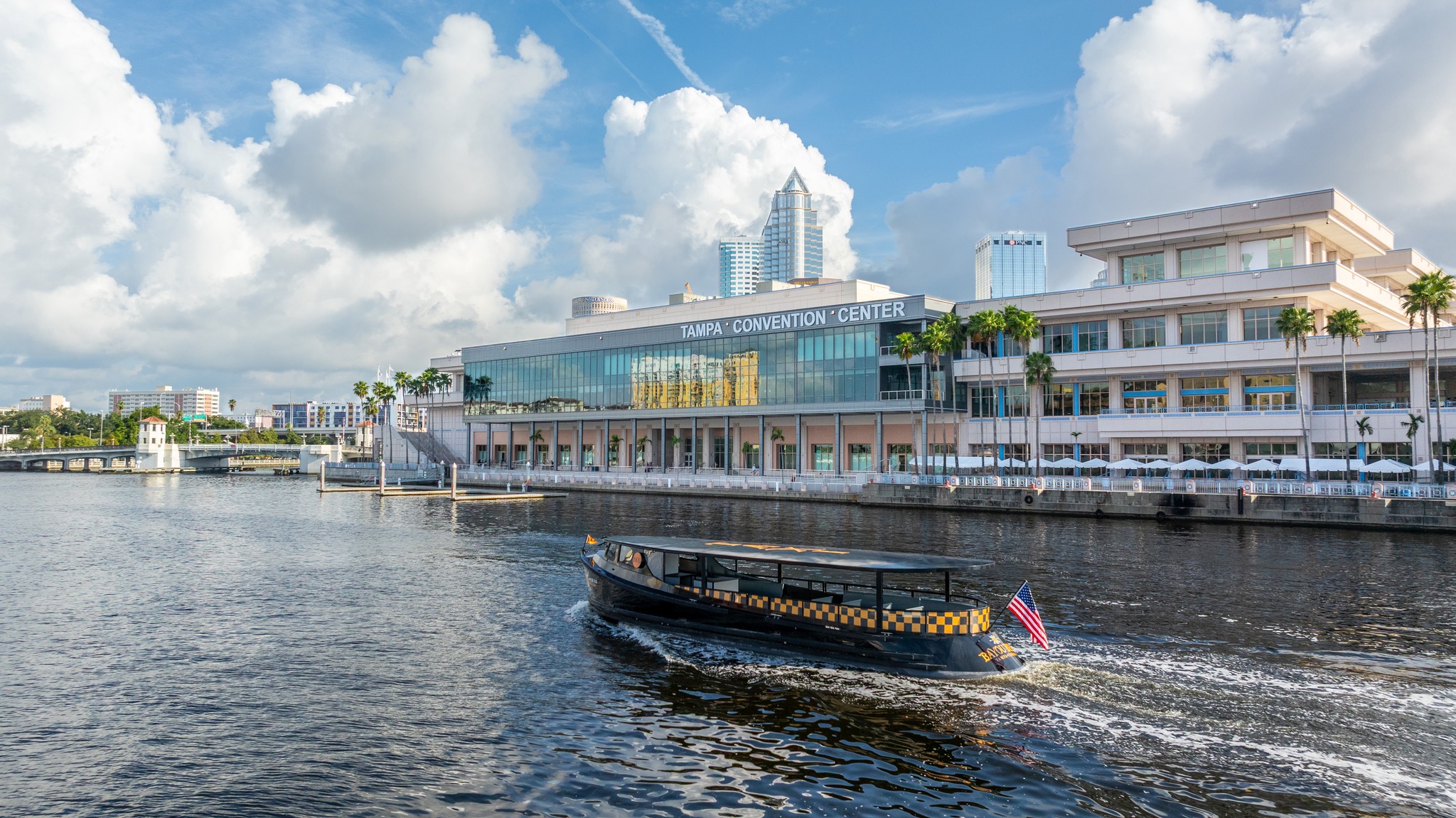 Exterior of Tampa Convention Center on the water with blue skies and city in the background and Pirate Water Taxi in the foreground.