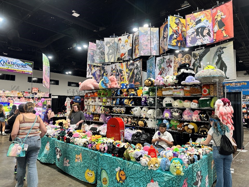 Merch booth in exhibit hall filled with stuffed animals and posters with attendees standing nearby.
