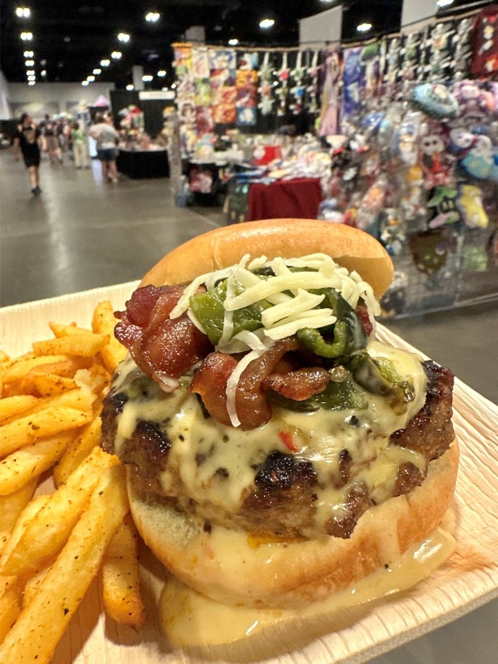 Burger loaded with cheese, bacon, and guacamole on plate with seasoned fries and exhibit hall vendors in the background.