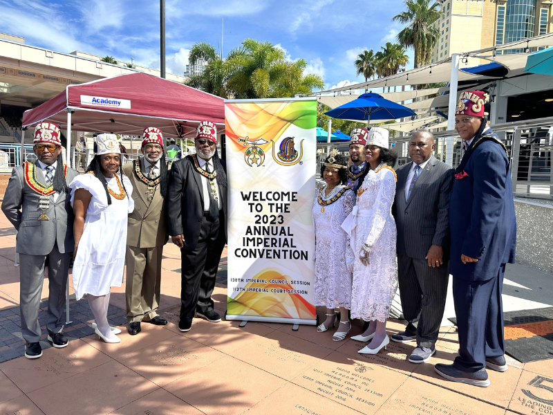 Group of Prince Hall Shriners members in traditional Shriners outfits gather around a sign that reads Welcome to the 2023 annual imperial convention in front of a plaque carved into the Tampa Riverwalk.