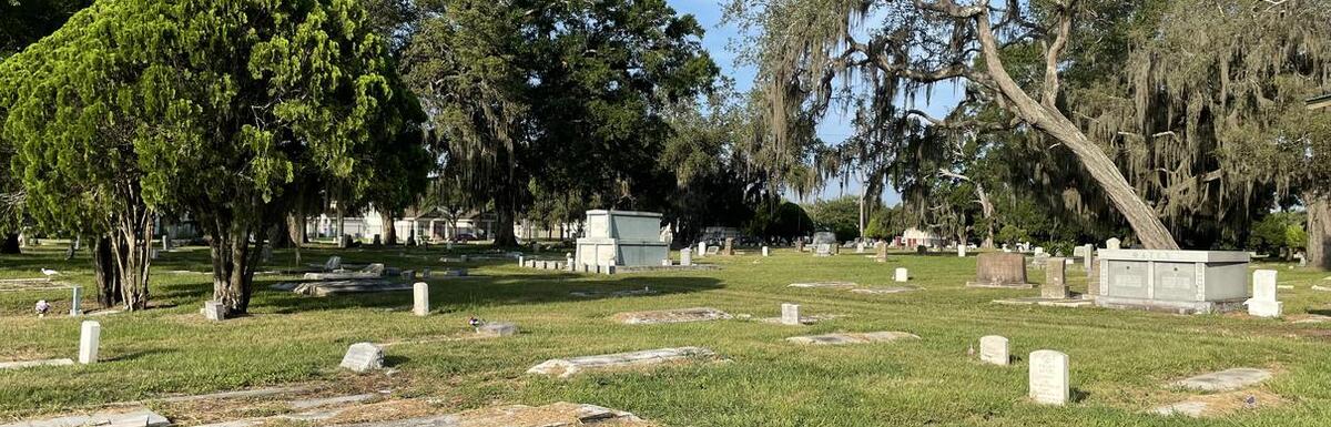 Grassy cemetery with scattered headstones and mature trees under a clear sky.