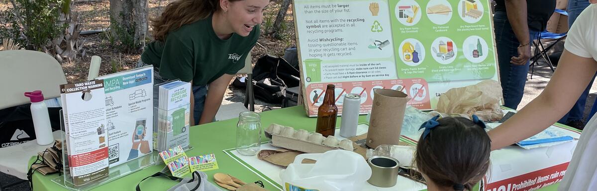 Outdoor event booth with educational display on recycling, featuring green tablecloth and visitors.