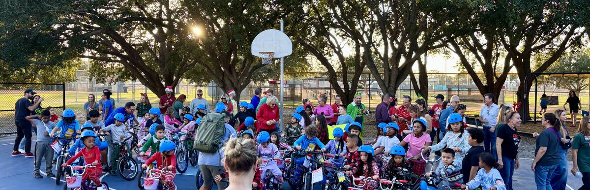 Large group of children with bikes and helmets gathered outdoors on a basketball court.
