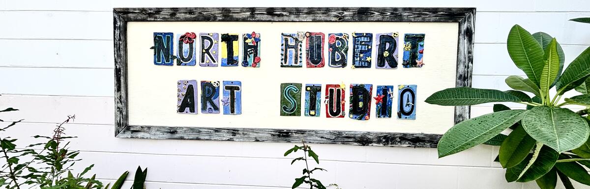 Sign reading "North Hubert Art Studio" on a white wall, framed by plants.