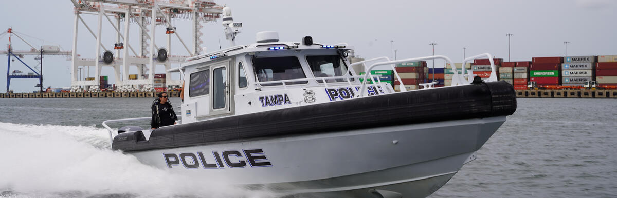 Police boat speeding on water with police helicopter flying above near shipping docks.