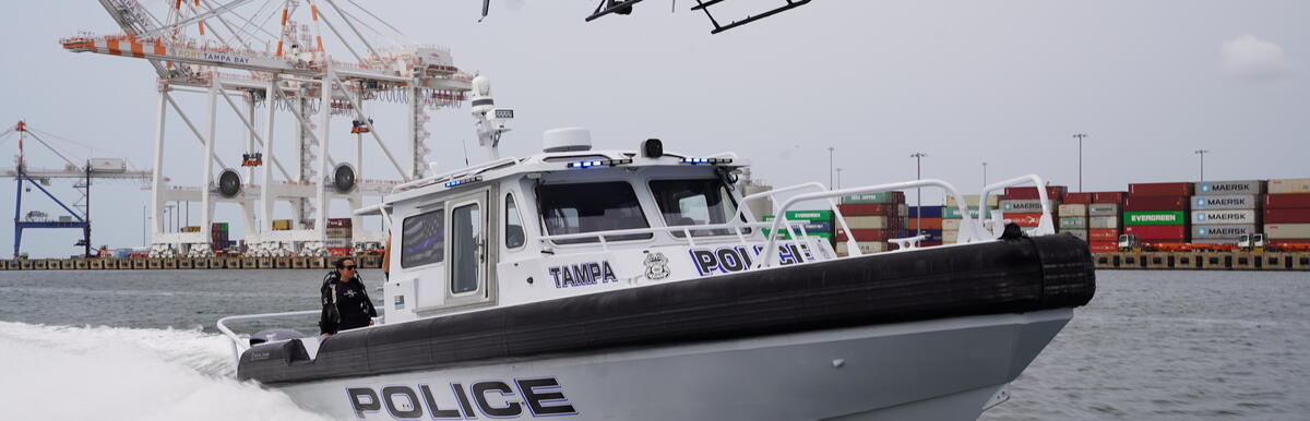 Police boat speeding on water with a police helicopter hovering above near shipping containers.