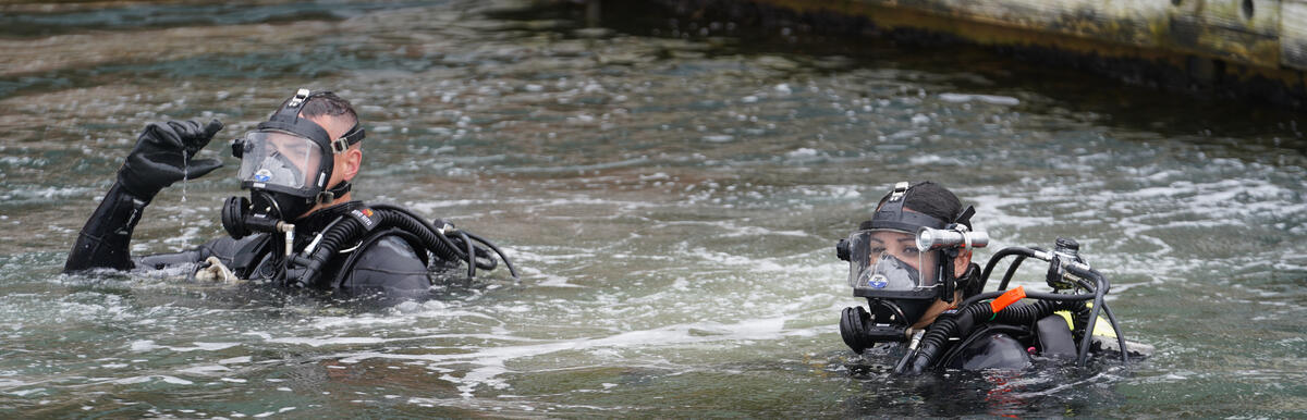 Two scuba divers in black wetsuits near a dock, one making a hand signal.