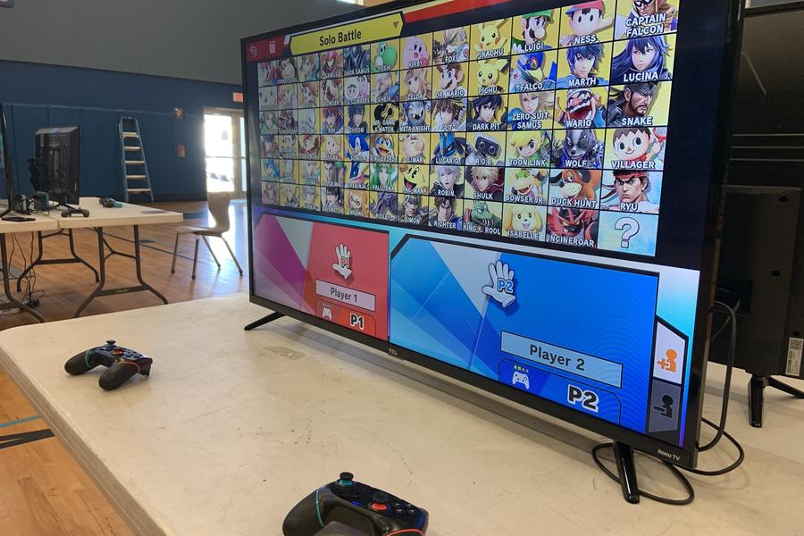 TV screen with two game controllers sitting on top of a table with Super Smash Bros. Ultimate character selection on screen
