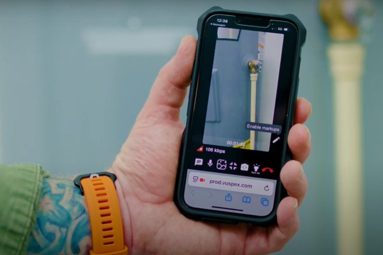 Hand holding a phone displaying a Vuspex inspection video with plumbing pipes.