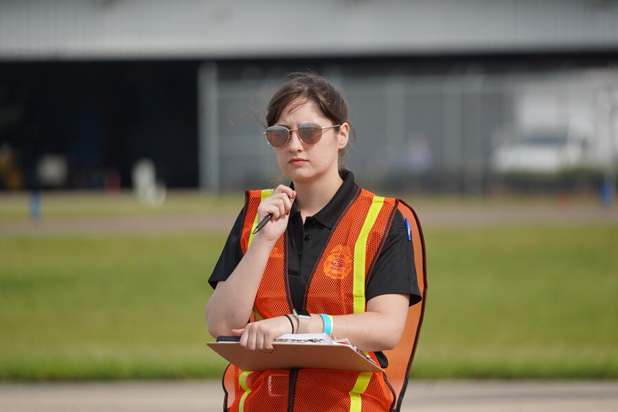 Woman in a safety vest with a clipboard, standing outdoors pensively.