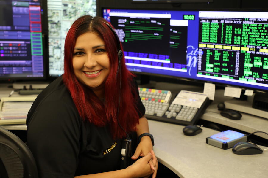 Woman smiling at a desk with multiple computer monitors in a control room.