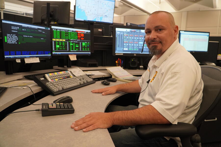Man sitting at a desk in a control room, multiple monitors display data and maps.