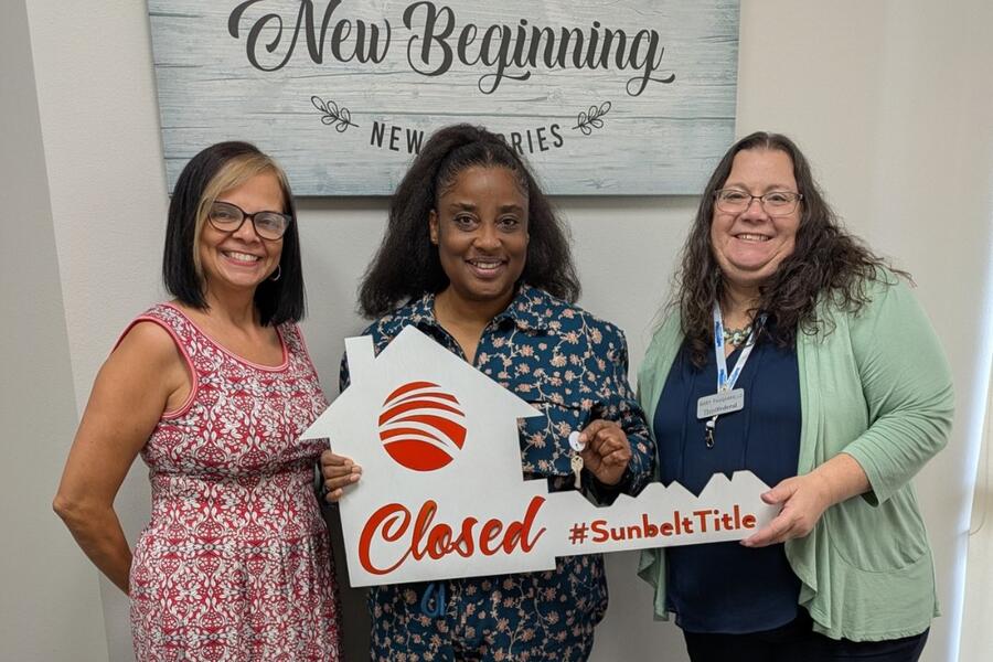 Three women smiling, one holds a "Closed" sign shaped like a house in an office setting.
