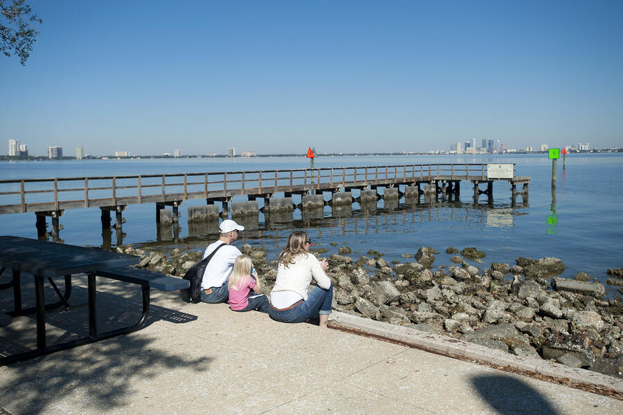 Coastal boardwalk with people sitting near rocky shore, city in background.