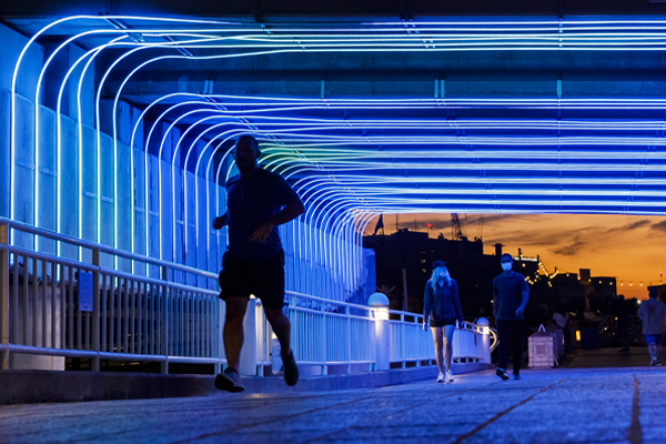 People walking and running under a bridge with blue bio-inspired LED light patterns at sunset.