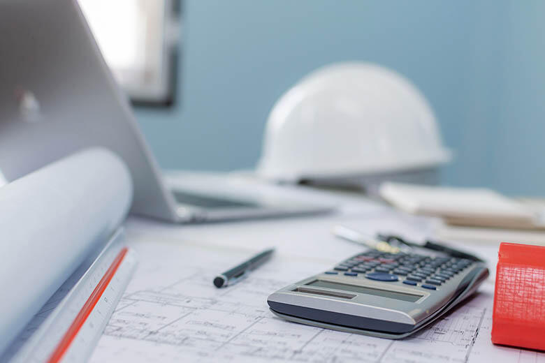 Blueprints, calculator, pen, laptop, and construction helmet on a desk in a workspace.