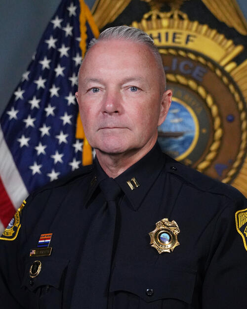 Captain in uniform with U.S. flag and police emblem in the background.