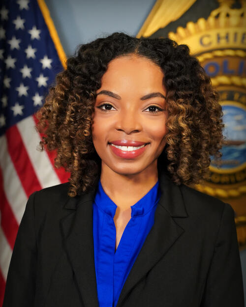 Smiling woman in professional attire in front of American flag and police emblem.