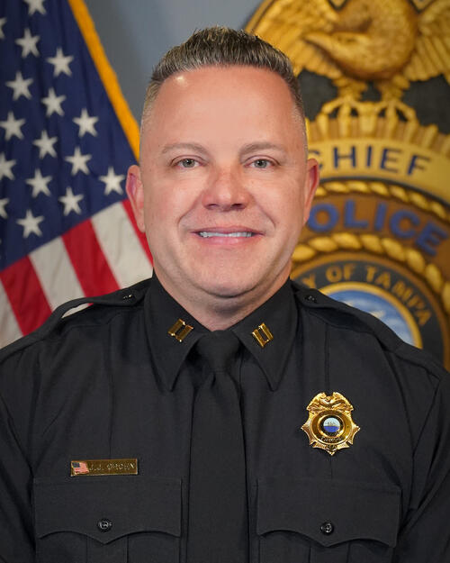 Police captain in uniform, standing in front of U.S. flag and police emblem.