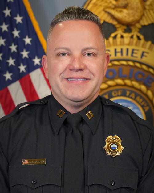 Police captain in uniform with a badge, flag, and police insignia in the background.