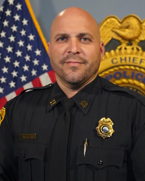 Police captain in uniform stands in front of a U.S. flag and police badge.