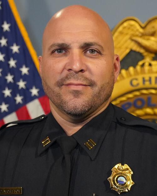 Police captain in uniform stands in front of a U.S. flag and police badge.