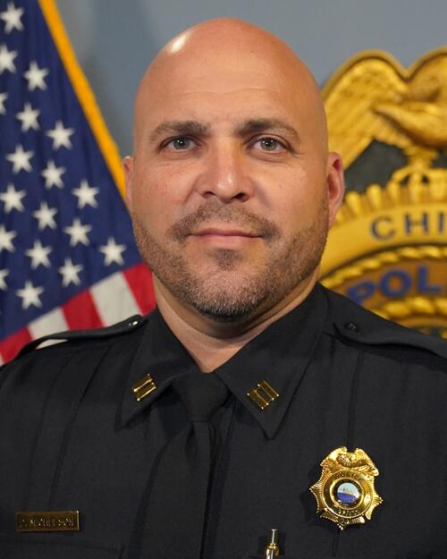 Police captain in uniform in front of a flag and badge display.