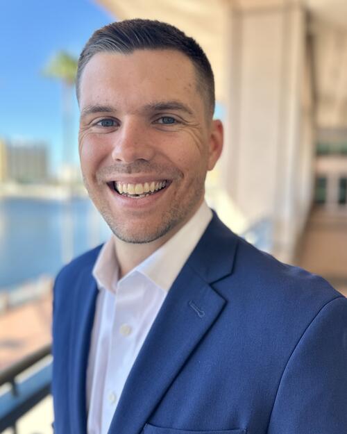 Smiling man in a blue suit on a sunny balcony with a waterfront view.