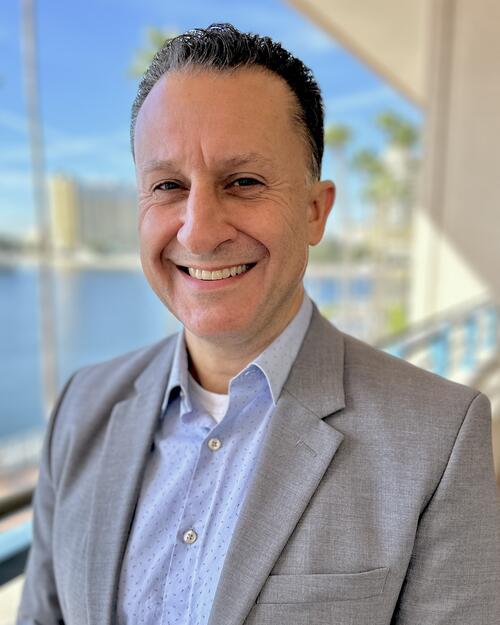 Smiling person in a gray blazer stands outside near water with a cityscape in the background.
