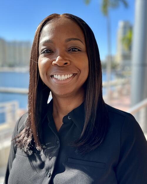 Smiling woman in a black shirt, standing near a waterfront, sunny day.