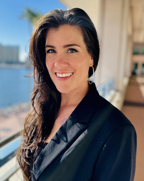Smiling woman in a black blazer, standing in a sunlit outdoor corridor.