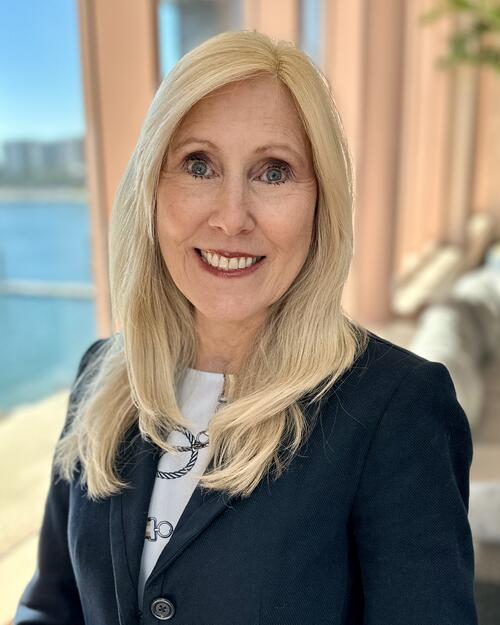Blonde woman smiling in a sunlit room with large windows overlooking water.