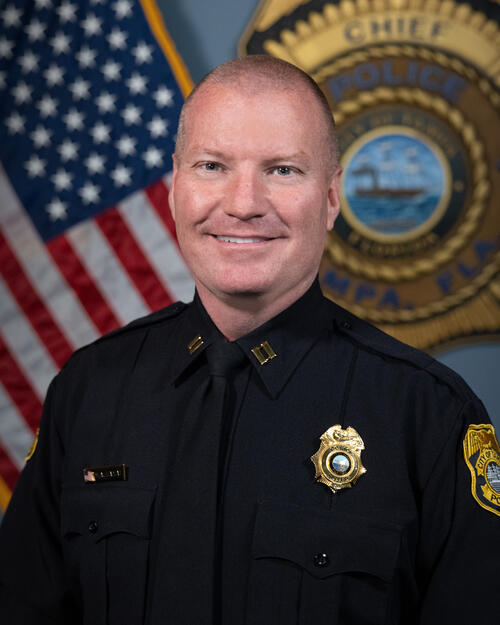Captain Kamat in uniform smiling, with a U.S. flag and police badge in the background.