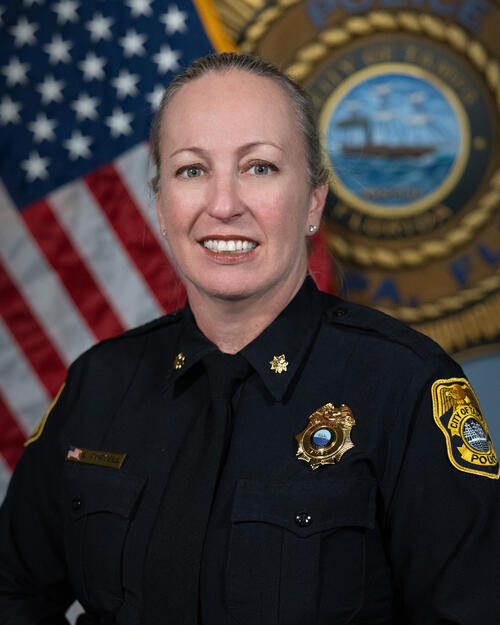 Major Purcell smiling, in uniform, against American and departmental flags backdrop.