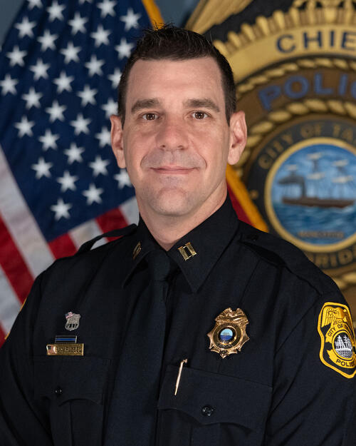 Police Captain Russel in dark uniform standing in front of U.S. flag and police emblem.