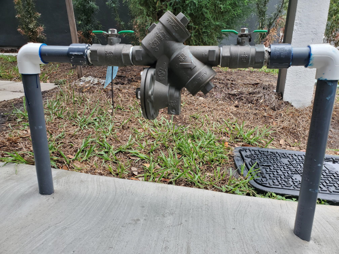 Backflow prevention device installed on outdoor plumbing with valves and PVC pipes.