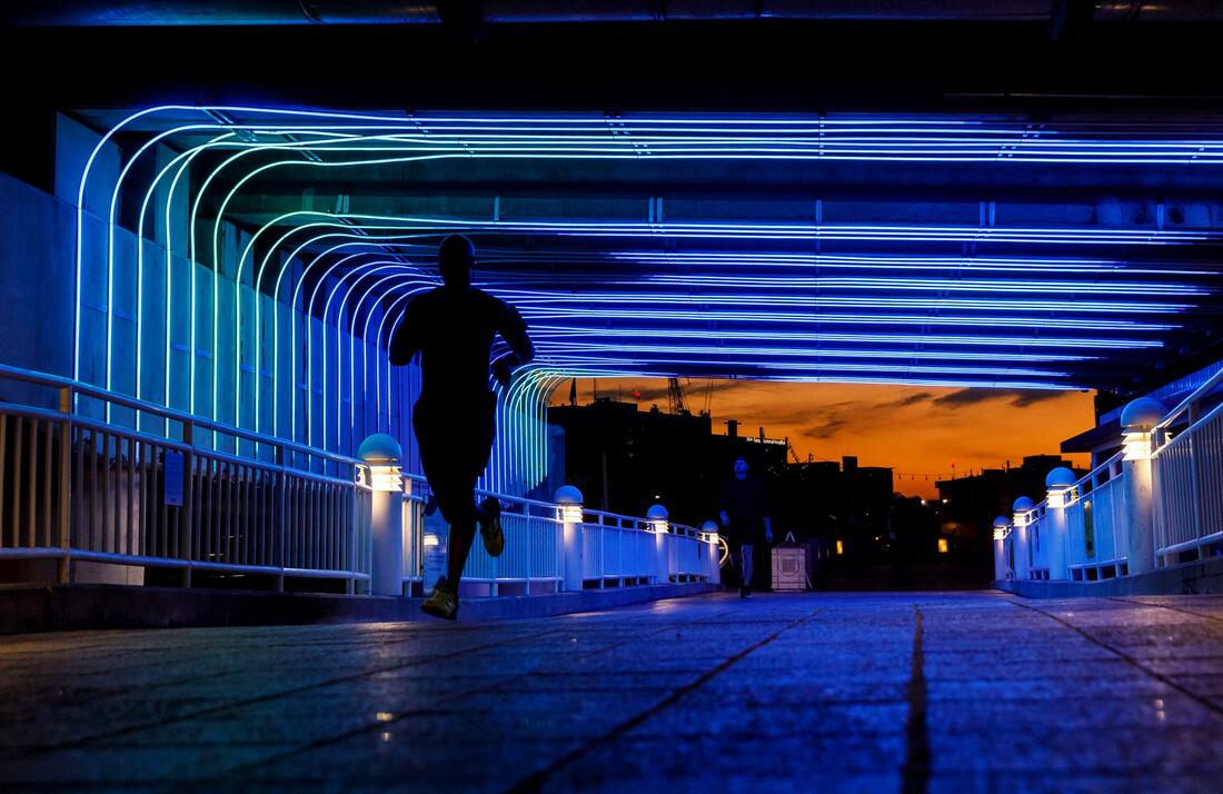Person running on a lit BioBridge by Andrea Polli at dusk, glowing blue lights overhead.