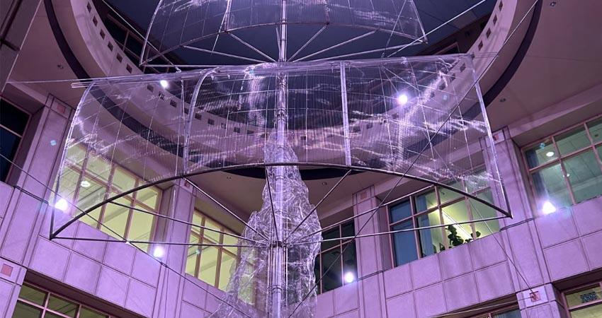 Large transparent sculpture resembling rising smoke, suspended indoors under purple lighting.