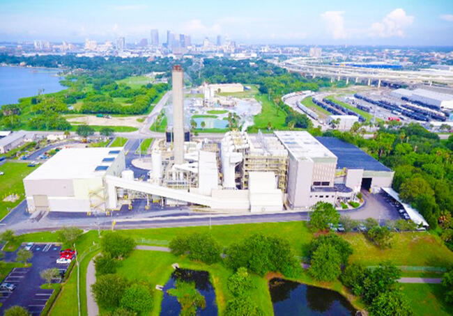 Aerial view of a waste-to-energy plant surrounded by greenery and distant cityscape.
