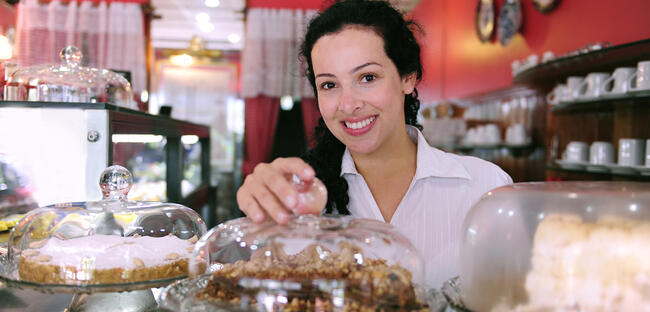 Smiling woman in a small bakery, displaying pastries under glass domes.