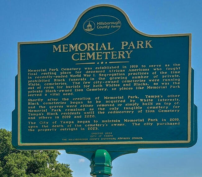 Memorial Park Cemetery sign with historical information in a grassy area, blue sky backdrop.
