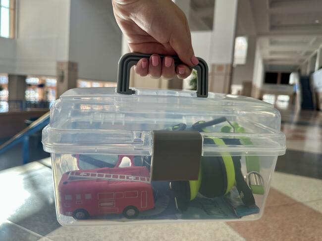 Clear plastic box with toy vehicles inside, held by a hand in a sunlit room.