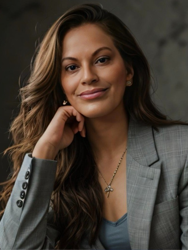 Smiling woman in a gray suit jacket, long wavy hair, against a dark background.