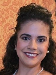 Smiling woman with curly hair and pearl jewelry, standing indoors.