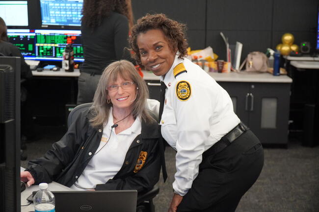 Two smiling women in uniform at a control room desk with screens.